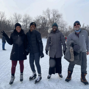 Three women wearing skates and one man wearing winter boots standing on the ice. All are smiling.